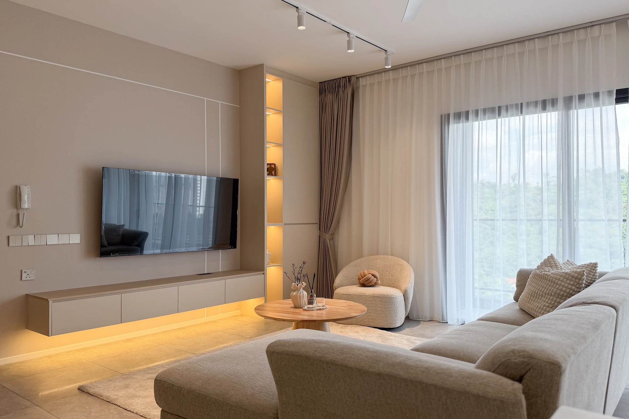 The Muji Apt living room in Allevia, Kuala Lumpur featuring a neutral sectional sofa, built-in TV console, warm LED lighting and floor-to-ceiling curtains.
