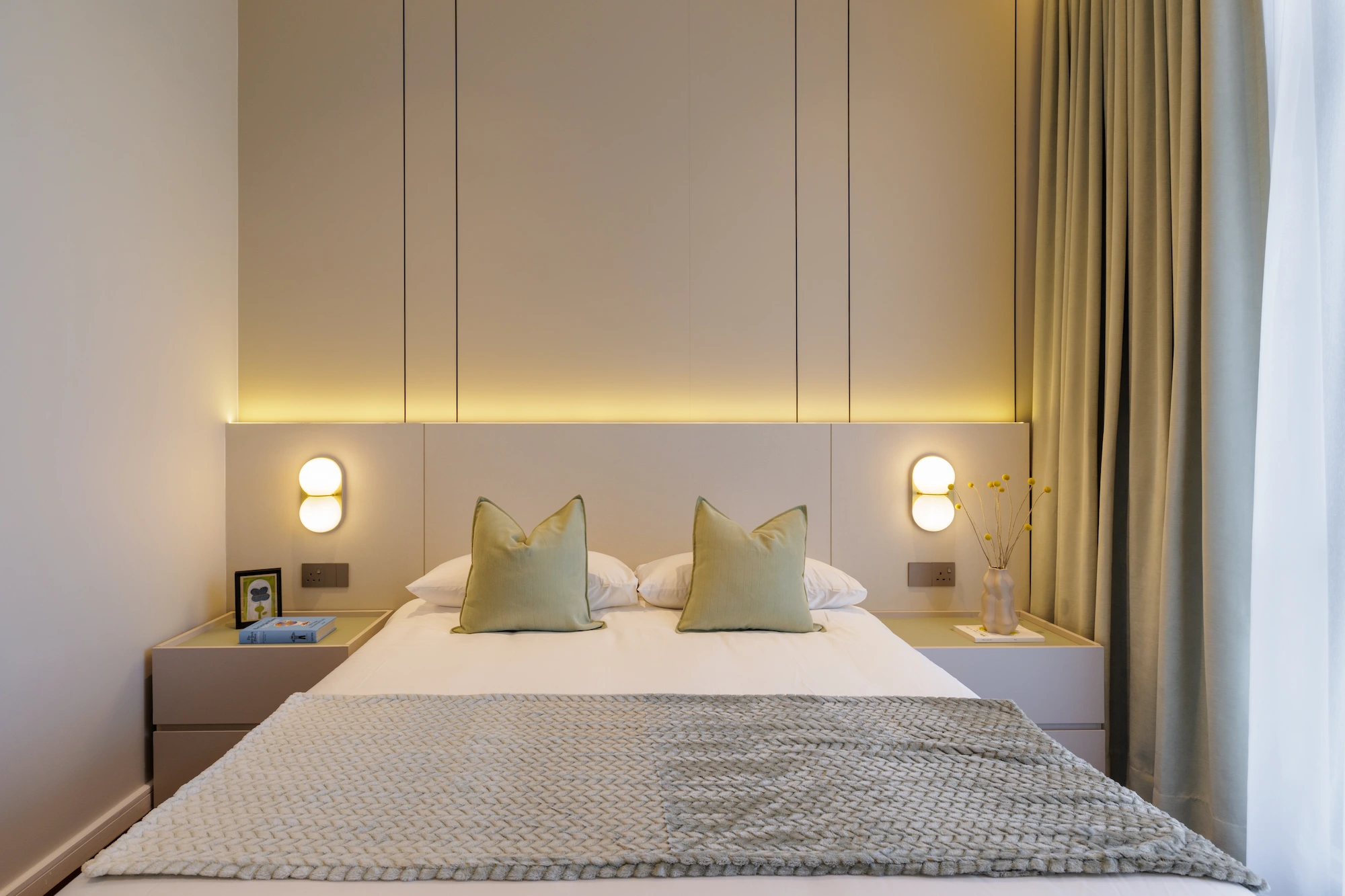 The Radius bedroom featuring a custom upholstered headboard, integrated wall lighting, green accent cushions, and warm wood flooring.