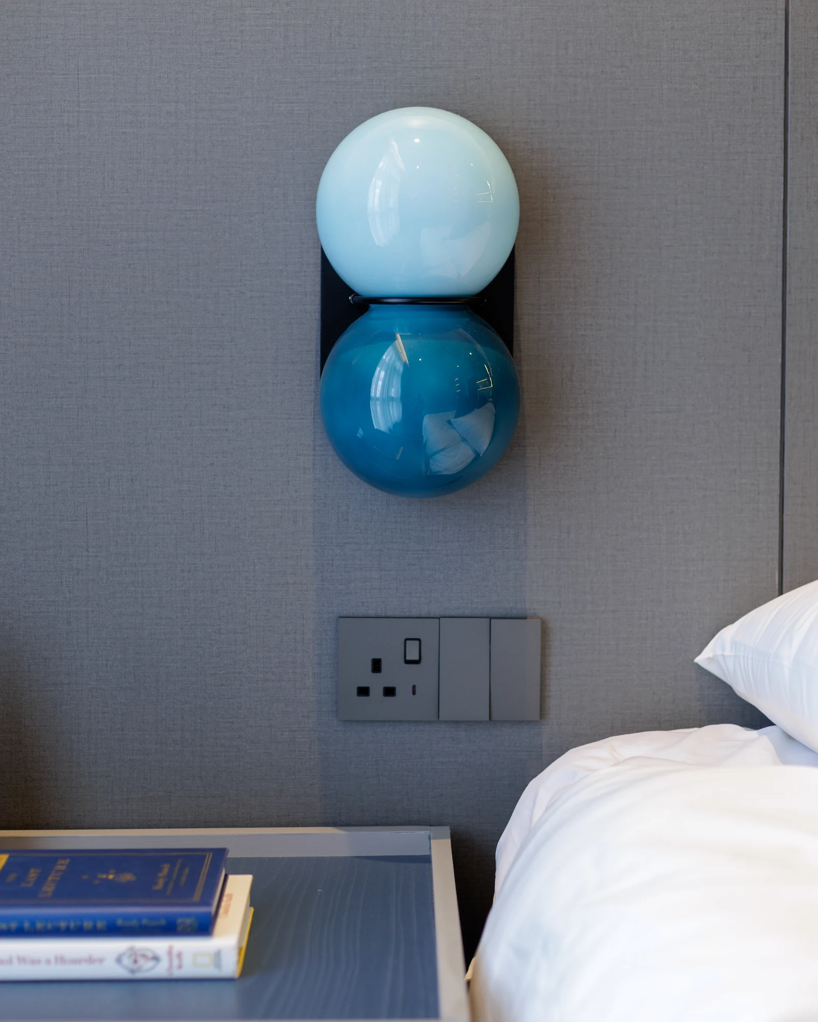 The Radius bedroom featuring a sculptural blue wall light, textured grey headboard, and styled bedside table with art books.