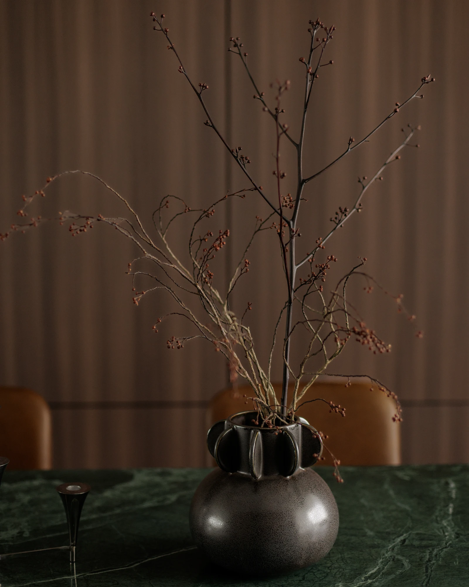 The Radius dining table styled with a sculptural dark vase, delicate branches, and a green marble tabletop against a dark wood wall.